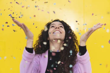 Cheerful woman playing with confetti against yellow background - MGRF01109
