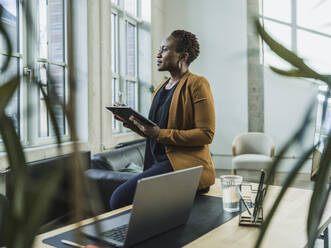 Thoughtful businesswoman holding tablet PC sitting at desk in office - MFF09551