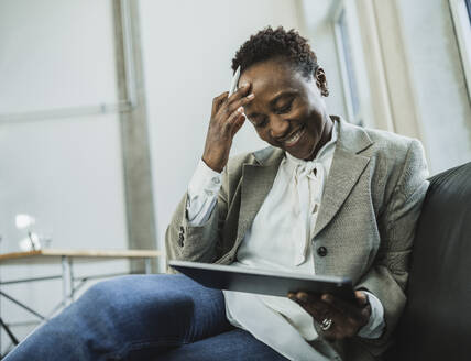 Smiling businesswoman with head in hand holding tablet PC on sofa at office - MFF09564