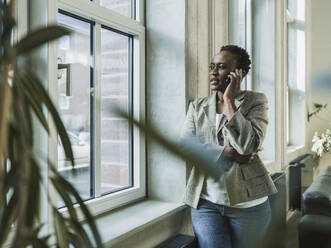 Businesswoman talking on smart phone leaning near window at office - MFF09574