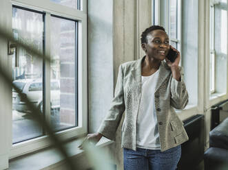 Smiling businesswoman talking on smart phone near window at office - MFF09575