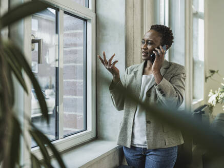 Geschäftsfrau gestikuliert und spricht auf Smartphone in der Nähe von Fenster im Büro - MFF09576