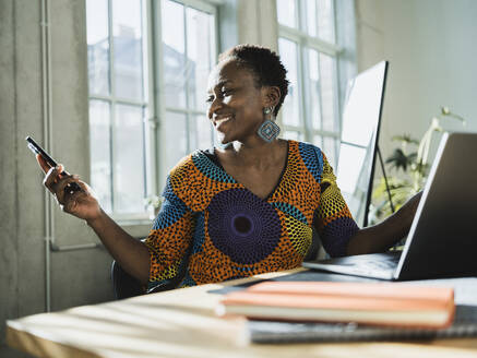 Lächelnde Geschäftsfrau, die ein Smartphone benutzt und am Schreibtisch im Büro sitzt - MFF09592
