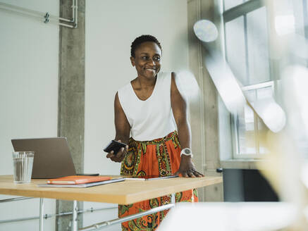 Lächelnde Geschäftsfrau, die ein Smartphone hält und sich an einen Schreibtisch im Büro lehnt - MFF09596