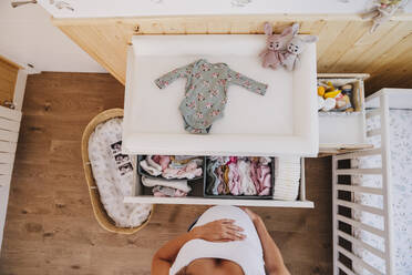 Pregnant woman standing near baby clothes at home - EBBF08668