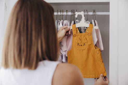 Frau hält Babykleidung in der Nähe des Kleiderschranks zu Hause - EBBF08671