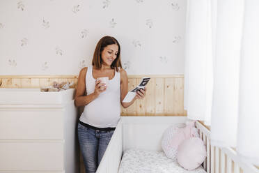 Smiling pregnant woman looking at ultrasound photographs near crib at home - EBBF08674