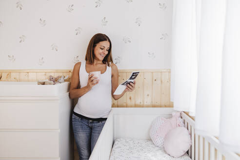 Lächelnde schwangere Frau betrachtet Ultraschallbilder in der Nähe der Krippe zu Hause - EBBF08674