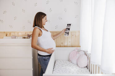 Schwangere Frau sieht sich Ultraschallbilder in der Nähe des Kinderbettes zu Hause an - EBBF08676