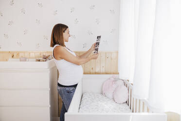 Pregnant woman looking at ultrasound photograph in nursery bedroom at home - EBBF08677