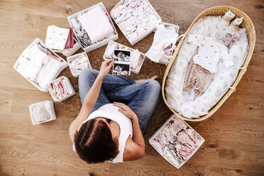 Pregnant woman holding ultrasound photographs near moses basket at home - EBBF08685