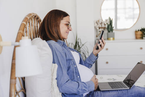 Lächelnde schwangere Freiberuflerin, die ihr Smartphone in der Nähe ihres Laptops im Home Office benutzt - EBBF08704