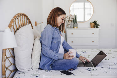 Pregnant freelancer holding mug and using laptop on bed at home office - EBBF08710