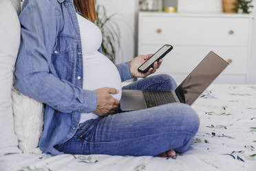 Pregnant woman sitting on bed with smart phone and laptop at home office - EBBF08711