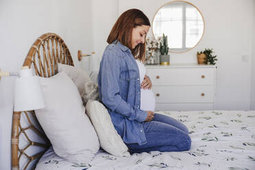 Schwangere Frau sitzt zu Hause auf dem Bett - EBBF08712