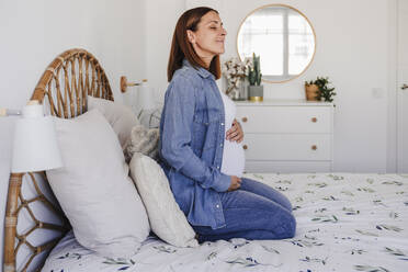 Pregnant woman with eyes closed sitting on bed at home - EBBF08713