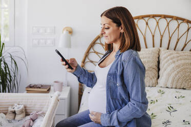 Schwangere Frau sitzt auf dem Bett und benutzt ihr Smartphone zu Hause - EBBF08716