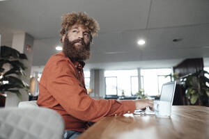 Mature businessman sitting with laptop at table in office cafeteria - JOSEF23432