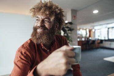 Lächelnder reifer Geschäftsmann mit Afrofrisur, der eine Kaffeetasse in einer Büro-Cafeteria hält - JOSEF23434