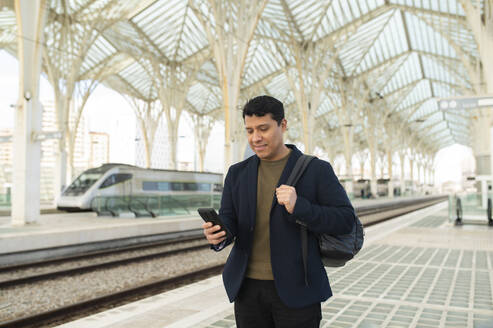 Lächelnder Geschäftsmann mit Smartphone auf dem Bahnsteig - VRAF00458