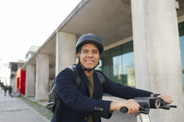 Smiling businessman wearing helmet and riding electric push scooter on sidewalk - VRAF00463