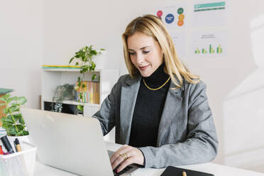 Smiling blond businesswoman working on laptop in office - XLGF03330