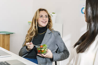 Glückliche Geschäftsfrau sitzt mit einer Schüssel Salat neben einem Kollegen am Schreibtisch - XLGF03346