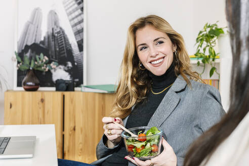 Lächelnde Geschäftsfrau sitzt mit einer Schüssel Salat neben einem Kollegen am Schreibtisch - XLGF03347