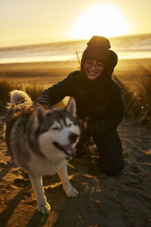 Lächelnde Frau kniend und Husky-Hund am Strand streichelnd - ANNF00831