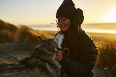 Lächelnde Frau streichelt Husky-Hund am Strand - ANNF00832