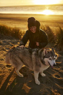 Lächelnde junge Frau streichelt Husky-Hund auf Sand am Strand - ANNF00833