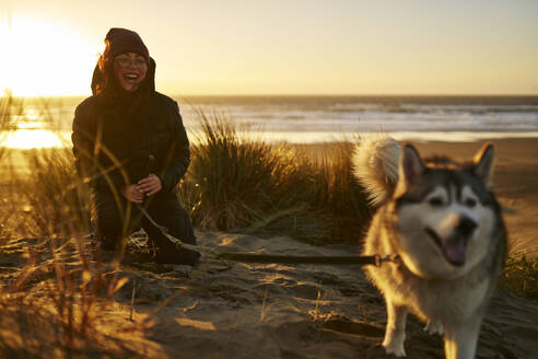 Fröhliche junge Frau spielt mit Husky-Hund am Strand - ANNF00836