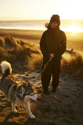 Glückliche junge Frau, die mit ihrem Husky-Hund am Strand spazieren geht - ANNF00840