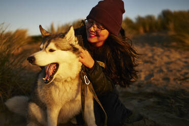 Smiling woman wearing knit hat and petting Husky dog at sunset - ANNF00842