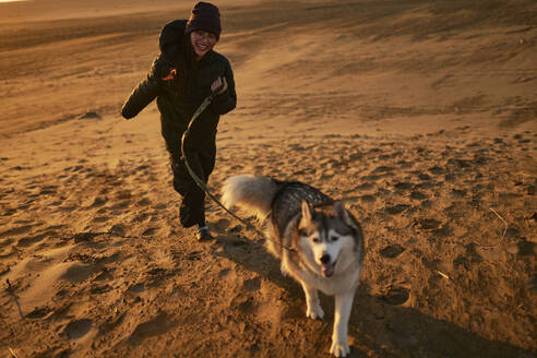 Fröhliche Frau läuft mit Hund auf Sand bei Sonnenuntergang - ANNF00846