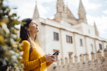 Smiling woman holding smart phone in front of building in city - JOSEF23455