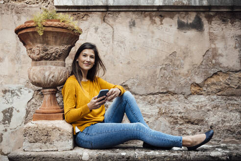 Lächelnde Frau hält Smartphone in der Nähe von Terrakotta-Pflanzentopf vor der Wand - JOSEF23469