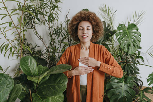 Smiling woman breathing with hands on chest near plants - YTF01833
