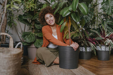 Lächelnde junge Frau sitzt mit Pflanzen auf einem Teppich - YTF01836