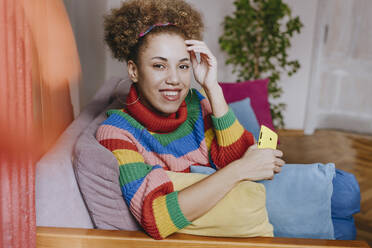 Happy young woman sitting with smart phone on sofa - YTF01846