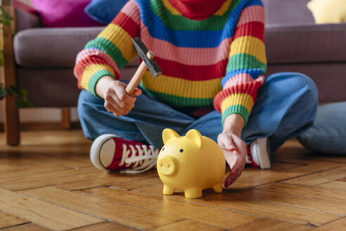 Woman sitting with hammer and piggy bank at home - YTF01857