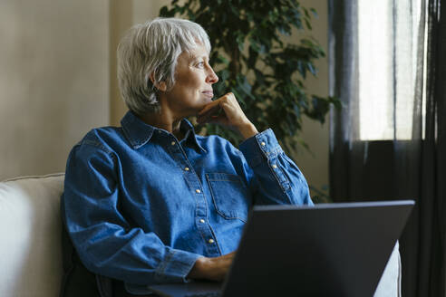 Thoughtful mature freelancer sitting with laptop at home office - EBSF04368
