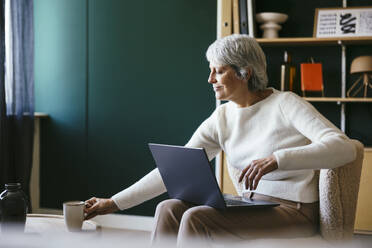 Freelancer sitting with laptop and picking up coffee mug at home office - EBSF04380