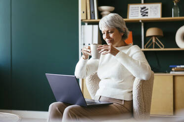 Älterer Freiberufler mit Kaffee und Blick auf den Laptop im Büro zu Hause - EBSF04381