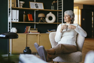 Smiling woman holding coffee mug and sitting on chair at home - EBSF04384