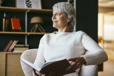 Smiling mature woman sitting and holding book at home - EBSF04386