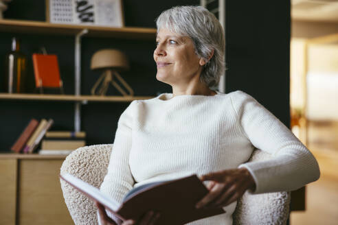 Lächelnde reife Frau sitzt und hält ein Buch zu Hause - EBSF04386