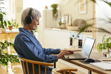 Smiling freelancer wearing wireless headphones and working on laptop at home office - EBSF04387