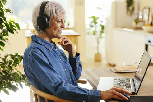 Smiling freelancer sitting with hand on chin using laptop at home office - EBSF04390