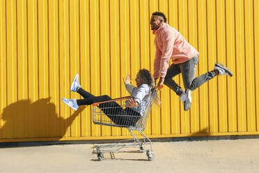 Carefree woman sitting in shopping cart near man jumping in front of yellow striped wall - SYEF00532